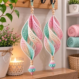 May include: Two hanging spiral ornaments crafted from crocheted fabric in pastel shades of pink, cream, and mint green. Each piece features a central wooden bead at the top and a small, rounded crocheted tassel at the bottom with wooden accents.