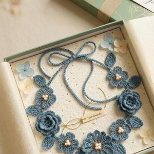 Può includere: Una collana di fiori all'uncinetto blu fatta a mano con dettagli di perle, presentata in una confezione regalo. La collana presenta un delicato design floreale ed è legata con un cordoncino blu. La confezione regalo è verde.