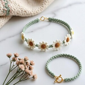 May include: A daisy chain necklace and bracelet set. The necklace is light green with white and yellow daisy accents. The bracelet is light green with a gold clasp. A sprig of pink flowers is in the lower left corner.