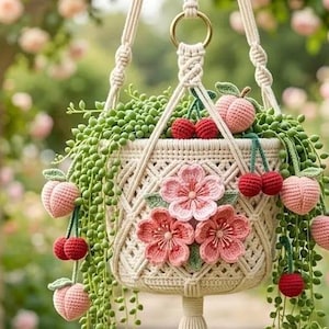 May include: A macrame hanging planter with a cream-colored woven design, adorned with crochet peaches, cherries, and pink flowers. The planter holds green trailing plants. The background features a blurred garden with pink roses.