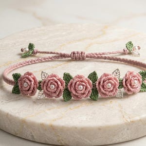 May include: A pink crocheted bracelet with five rose flowers, each with a pearl centre and green leaves. The bracelet has a sliding knot closure and silver-coloured leaf accents. The bracelet is displayed on a marble surface.