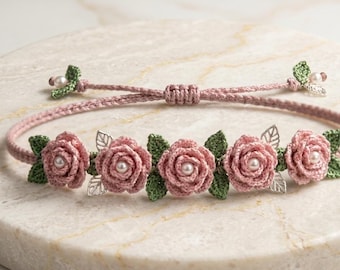 Pode incluir: Uma pulseira rosa de crochê com cinco flores de rosa com centros de pérolas e folhas verdes. A pulseira tem um fecho de nó deslizante e detalhes de folhas prateadas. A pulseira é exibida em uma superfície de mármore.
