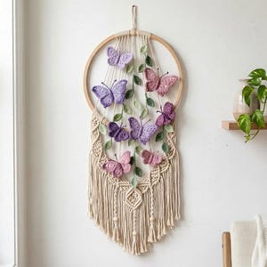 May include: A macrame wall hanging featuring a wooden hoop, adorned with cream-colored knotted threads, and embellished with purple and pink butterflies and green leaves. The wall decor is designed to be hung on a wall.