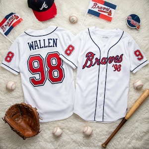 以下が含まれることがあります： 赤と青のアクセントが付いた2つの白い野球のユニフォーム。 「Braves」の文字と「98」と「8」の数字が特徴です。 野球帽、野球ボール、野球グローブ、バットも写っています。