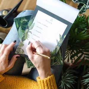 May include: A person writing on a notepad with a green and black tropical leaf design. The notepad has the word "REGAR" at the top and a list of plants to water, including "Limes & Verbena", "Avocado", "Monstera", "Heucheras", and "Ficus".