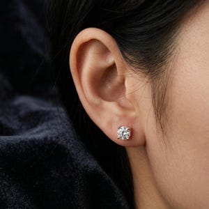May include: Close-up of a sparkling, round-cut diamond stud earring set in a rose gold setting. The earring is worn on an ear, showcasing its brilliance and design. The background is dark, emphasizing the earring's sparkle.