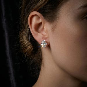 Puede incluir: Primer plano de la oreja de una mujer con un gran pendiente de diamante redondo y brillante. El pendiente está engastado en un engaste de oro o platino. El fondo es oscuro, enfatizando el brillo del diamante.