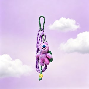Op de afbeelding: Een paarse en roze tie-dye vilten berenhanger bevestigd aan een paars touwkoord met een groene karabijnhaak. De beer heeft een donkerblauwe neus en een gestikte glimlach. Een kleine gele kraal met een zwart smiley-gezicht zit onderaan.