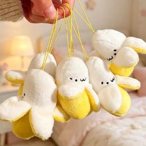 Op de afbeelding: Een tros pluche bananenhangers van zachte witte en gele stof, elk met een eenvoudig geborduurd gezicht bestaande uit twee zwarte ogen en een gebogen mond. Elke pluche banaan is bevestigd aan een dun geel koordje om op te hangen.