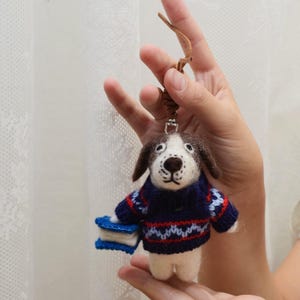 May include: A needle-felted dog ornament featuring a white and brown face with dark spots, wearing a navy blue, red, and light blue patterned knit sweater. The dog holds a small blue and white book-shaped object and hangs from a brown cord.