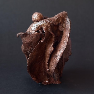May include: A dark brown ceramic sculpture with a rough, textured surface. The object features a spherical top element resting above a series of vertical, folded, and undulating clay layers that create a complex, organic, and abstract form.