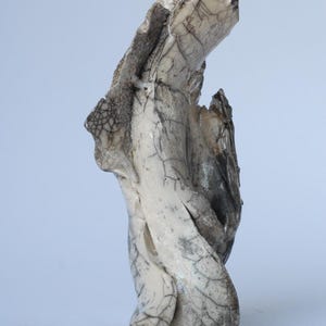 May include: Abstract ceramic sculpture with a textured, organic form. The sculpture features a blend of off-white, gray, and black tones, with intricate crackle patterns across its surface. The piece has a unique, twisted shape.