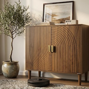May include: A wooden cabinet with a carved design and brass handles. The cabinet is set against a wall with a framed landscape painting and a small radio. A potted olive tree and a robotic vacuum cleaner are in the foreground.