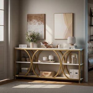 May include: A long console table with a gold metal frame featuring large circular accents and white shelves. The table holds various decorative items including a plant, candles, books, and storage boxes, with two framed art prints hanging above it.