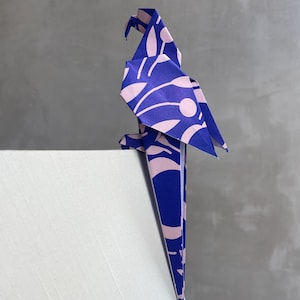 Può includere: Un pappagallo origami blu e rosa appollaiato su una superficie bianca. L'uccello di carta presenta un motivo ripetuto di forme rosa a forma di foglia su sfondo blu. Il becco e la coda del pappagallo sono appuntiti.