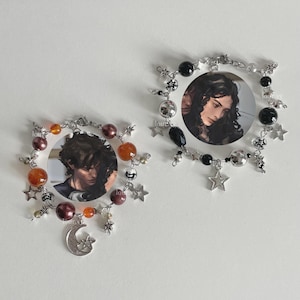 May include: Two round, silver-toned charm bracelets. Each bracelet features a circular photo frame with a portrait. The bracelets are adorned with beads in various colors, star charms, and a crescent moon charm.