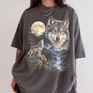 Pode incluir: Uma t-shirt de manga curta em cinzento-carvão com uma impressão gráfica de uma cabeça de lobo grande, um lobo mais pequeno a uivar para a lua cheia, uma cordilheira, pinheiros e uma cascata que desagua num rio.