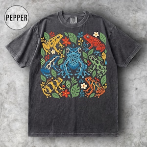May include: A charcoal grey short-sleeve t-shirt featuring a central graphic print of five colourful frogs in yellow, blue, red, green, and orange, surrounded by various green leaves, small red berries, and white flowers. The text PEPPER is visible.