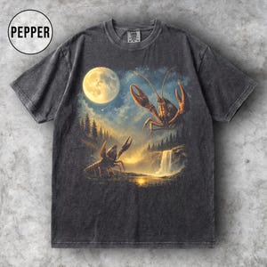 Puede incluir: Camiseta gris oscuro con un paisaje surrealista con dos cangrejos de río, una luna llena y una cascada. El gráfico tiene un aspecto vintage y desgastado. La palabra "PEPPER" está en un círculo en la parte superior izquierda.