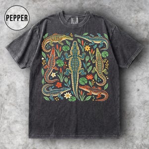 Könnte beinhalten: Ein anthrazitfarbenes Kurzarm-T-Shirt mit einer zentralen Grafik aus mehreren bunten Alligatoren, umgeben von verschiedenen Wasserpflanzen und Blumen. Das T-Shirt hat einen Rundhalsausschnitt und ein Etikett im Nackenbereich mit dem Text PEPPER.