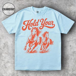 May include: Light blue t-shirt with the text "Hold Your" in red script above a graphic of two running horses in red. The shirt has short sleeves and a crew neck.
