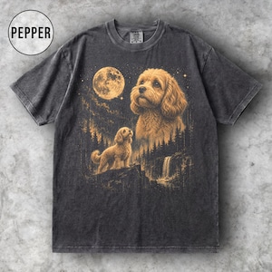 Op de afbeelding: Een antracietgrijs t-shirt met korte mouwen en een grafische print van twee goudkleurige honden in een boslandschap onder een volle maan. Het ontwerp bevat een waterval, dennenbomen en het woord PEPPER in een cirkel in de linkerbovenhoek.