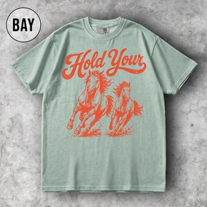 May include: A light green t-shirt featuring a distressed orange graphic of two running horses and the words "Hold Your" in a vintage font. The shirt has a classic crew neck and short sleeves.