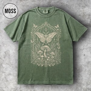 May include: A moss green t-shirt with a detailed cream-colored graphic featuring a moth, mushrooms, and botanical elements. The word "MOSS" is in a black circle in the upper left corner. The shirt has a classic crew neck and short sleeves.