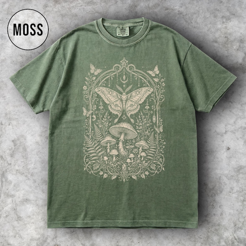 May include: A moss green t-shirt with a detailed cream-colored graphic featuring a moth, mushrooms, and botanical elements. The word "MOSS" is in a black circle in the upper left corner. The shirt has a classic crew neck and short sleeves.