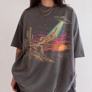 Może przedstawiać: Ciemnoszary t-shirt z krótkim rękawem z nadrukiem graficznym przedstawiającym złotego psa unoszonego przez promień UFO. Scena obejmuje pustynny krajobraz z kaktusem, skalistym klifem, zachodem słońca i rozgwieżdżonym niebem w odcieniach pomarańczu i fioletu.