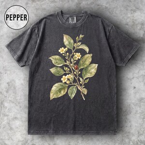 K&ouml;nnte beinhalten: Dunkelgraues T-Shirt mit einer botanischen Illustration eines bl&uuml;henden Zweigs mit gr&uuml;nen Bl&auml;ttern, gelben Bl&uuml;ten und einem roten Marienk&auml;fer. Das Wort "PEPPER" befindet sich in einem wei&szlig;en Kreis. Das Shirt hat einen Vintage-Look.
