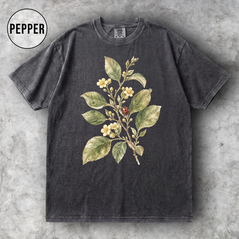 K&ouml;nnte beinhalten: Dunkelgraues T-Shirt mit einer botanischen Illustration eines bl&uuml;henden Zweigs mit gr&uuml;nen Bl&auml;ttern, gelben Bl&uuml;ten und einem roten Marienk&auml;fer. Das Wort "PEPPER" befindet sich in einem wei&szlig;en Kreis. Das Shirt hat einen Vintage-Look.
