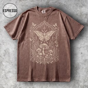 May include: Espresso-colored t-shirt featuring a detailed cream-colored graphic of a moth, mushrooms, and foliage within an ornate frame. The shirt has short sleeves and a crew neck. The word "ESPRESSO" is in a circle in the upper left corner.