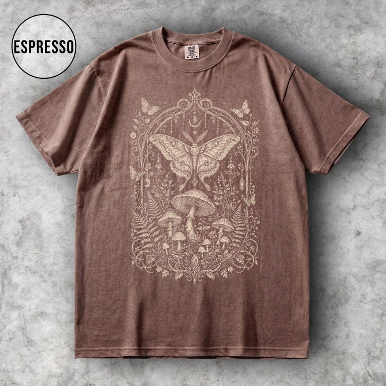 May include: Espresso-colored t-shirt featuring a detailed cream-colored graphic of a moth, mushrooms, and foliage within an ornate frame. The shirt has short sleeves and a crew neck. The word "ESPRESSO" is in a circle in the upper left corner.