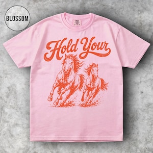 May include: A light pink t-shirt featuring the text "Hold Your" in a vintage style font above a graphic of two running horses in orange. The shirt has a crew neck and short sleeves.