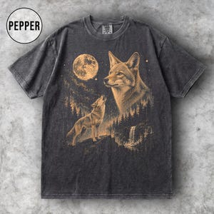 May include: Charcoal grey t-shirt with a detailed graphic of two coyotes under a full moon. One coyote is howling, and the other is looking towards the moon. The design includes a forest and waterfall. The word "PEPPER" is in a circle at the top left.