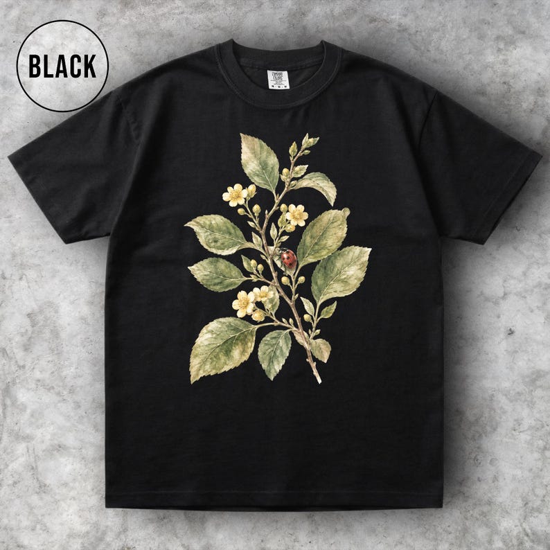 K&ouml;nnte beinhalten: Schwarzes T-Shirt mit botanischer Illustration. Das Design zeigt einen Zweig mit gr&uuml;nen Bl&auml;ttern, kleinen gelben Bl&uuml;ten und einem roten Marienk&auml;fer. Das Wort "BLACK" befindet sich in einem wei&szlig;en Kreis oben links.