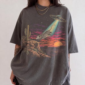 Puede incluir: Una camiseta gris carbón de manga corta con un estampado gráfico de una zarigüeya siendo abducida por un OVNI sobre un paisaje desértico con un cactus, terreno rocoso y un cielo al atardecer con tonos púrpuras y anaranjados.