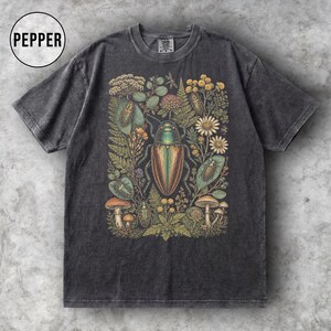 以下が含まれることがあります： チャコールグレーの半袖Tシャツ。中央には、大きな玉虫色の甲虫と、その周囲に描かれた野草、シダ、小さな昆虫、キノコなどの詳細な植物イラストが、アースカラーでグラフィックプリントされています。