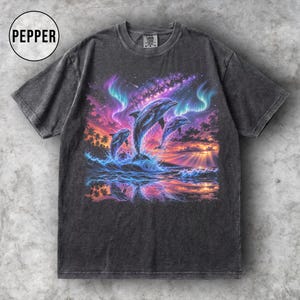 Peut inclure: T-shirt gris foncé avec un motif coloré de trois dauphins sautant hors des vagues de l'océan sous un coucher de soleil et des aurores boréales. Le t-shirt a un aspect vintage et délavé, et un col rond.