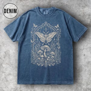 May include: Denim blue t-shirt featuring a detailed cream-colored graphic of a moth, mushrooms, and foliage within an arched frame. The word "DENIM" is in a white circle in the upper left corner. The shirt has short sleeves and a crew neck.