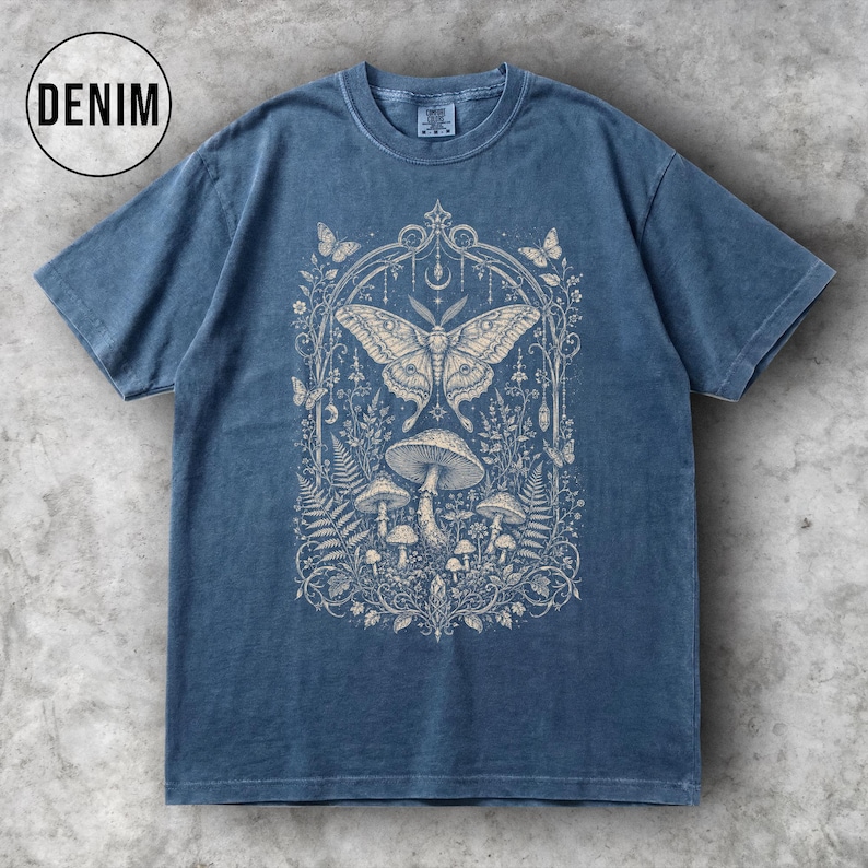 May include: Denim blue t-shirt featuring a detailed cream-colored graphic of a moth, mushrooms, and foliage within an arched frame. The word "DENIM" is in a white circle in the upper left corner. The shirt has short sleeves and a crew neck.