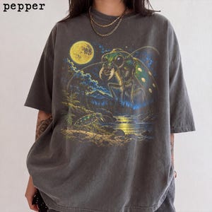 Puede incluir: Una camiseta gris carbón de manga corta con un estampado gráfico de un escarabajo verde grande en un paisaje forestal bajo una luna llena. La palabra pepper está impresa en la esquina superior izquierda. El diseño incluye un río y árboles.