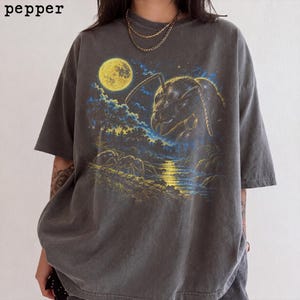 Può includere: Una t-shirt a maniche corte grigio antracite con una stampa grafica di una grande testa di formica che sovrasta un paesaggio boschivo con luna piena e un fiume. La parola pepper è stampata nell'angolo in alto a sinistra in carattere sans-serif.