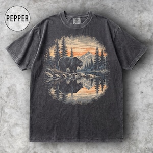以下が含まれることがあります： チャコールグレーのTシャツで、湖のそばに立つクマと、背景に松の木と山々が描かれた円形のグラフィックが特徴です。デザインには、水面に映るクマと風景の反射が含まれています。円の中に「PEPPER」という文字がプリントされています。
