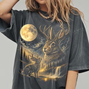 May include: Dark gray t-shirt featuring a detailed illustration of a jackalope with antlers, a full moon, and a waterfall scene. The design has a vintage, distressed look. The shirt is a relaxed fit.