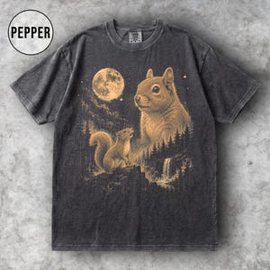 Puede incluir: Camiseta gris oscuro con una ilustración detallada de dos ardillas bajo una luna llena. El diseño incluye una cascada y elementos forestales, con la palabra "PEPPER" en un círculo en la parte superior izquierda.