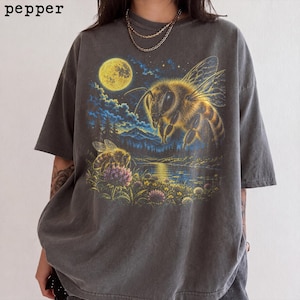 Può includere: Una t-shirt grigio antracite a maniche corte con una stampa grafica di una grande ape che vola sopra un'ape più piccola su un fiore vicino a un lago, montagne e una luna piena. La parola pepper è stampata nell'angolo in alto a sinistra.