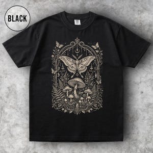 May include: Black t-shirt featuring a detailed beige illustration of a moth, mushrooms, and foliage within an ornate frame. The word "BLACK" is in a white circle in the upper left corner. The design is centered on the shirt.
