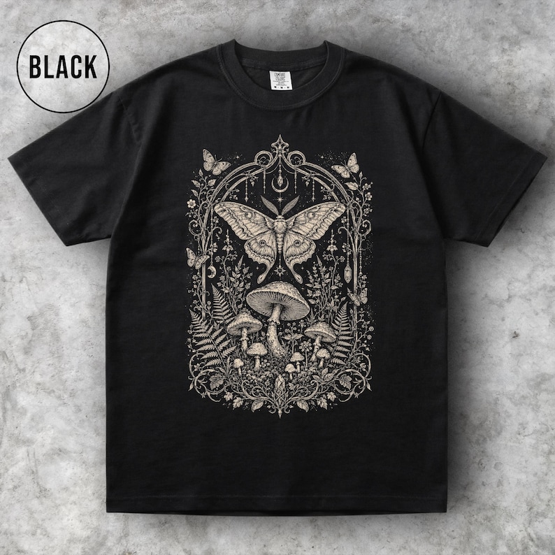 May include: Black t-shirt featuring a detailed beige illustration of a moth, mushrooms, and foliage within an ornate frame. The word "BLACK" is in a white circle in the upper left corner. The design is centered on the shirt.